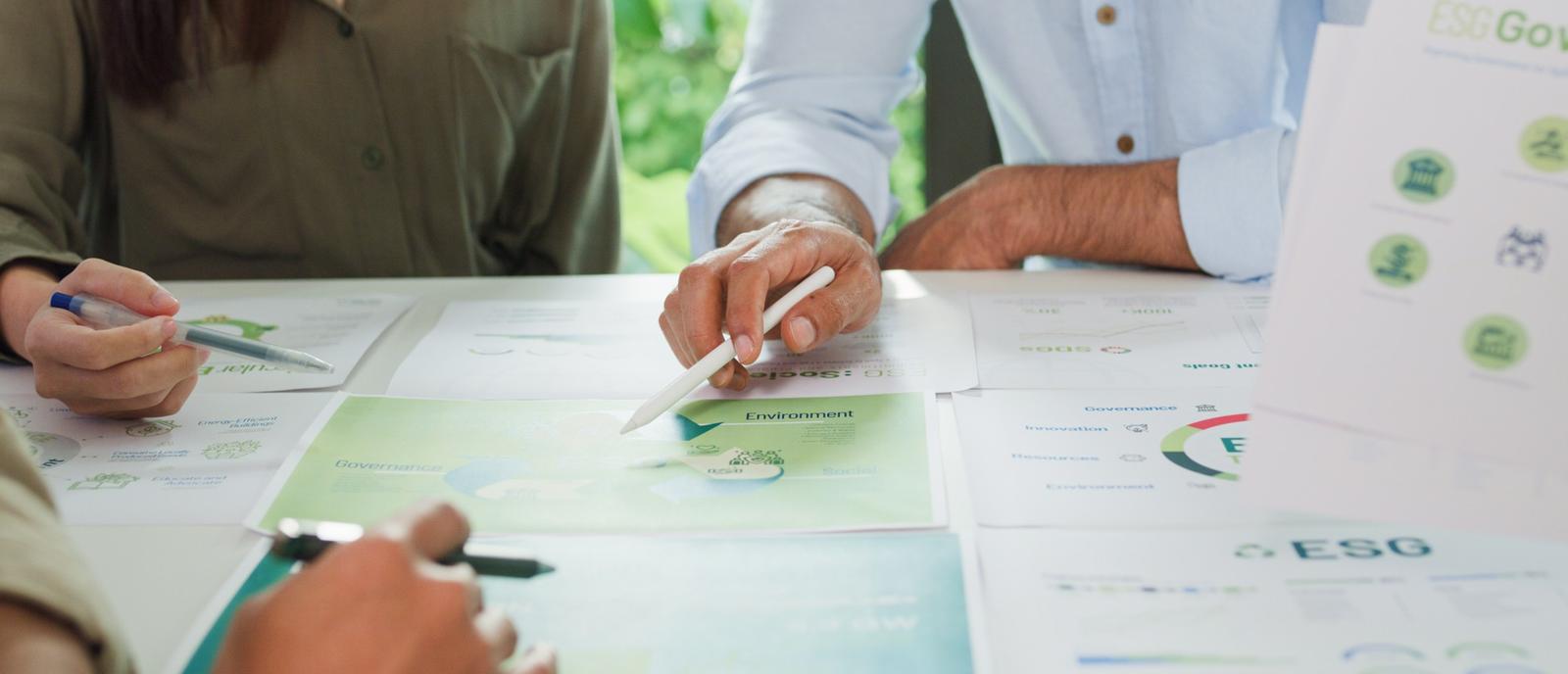 Auf einem Tisch liegen verschiedene Dokumente. Um den Tisch herum sitzen mehrere Personen. Zu sehen sind ihre Hände, die auf ein bestimmtes Blatt verweisen.