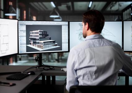 Zu sehen ist die Rückansicht eines Mannes, der an einem Schreibtisch vor mehreren Bildschirmen sitzt. Auf den Monitoren sind ein 3D-Modell sowie weitere Informationen zu einem Gebäude zu sehen.