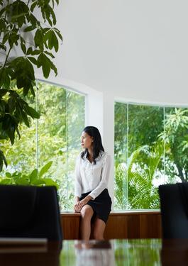 Eine schick gekleidete Frau sitzt seitlich auf der Fensterbank in einem Büro und blickt aus dem Fenster. Im Hintergrund befindet sich eine Fensterfront, durch die eine Fülle an grünen Pflanzen zu sehen ist.