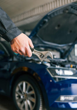 Foto auf dem eine Hand mit einem Schraubenschlüssel, in front eines Autos mit offener Motorhaube zusehen ist 