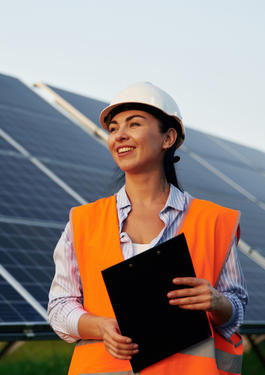 Frau in Arbeitsschutzkleidung hält ein Klemmbrett in den Händen. Im Hintergrund sind Solarpanele zu sehen.