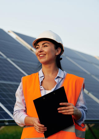 EIne Frau vor einer Solaranlage, in Arbeitsschutzkleidung und mit einem Tablet in der Hand