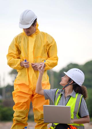 Zu sehen sind zwei Personen. Ein Mann in einem gelben Arbeitsschutzanzug hält ein Röhrchen zur Probenahme in der Hand. Vor ihm befindet sich eine Frau mit einem Laptop, die ihm ein weiteres Röhrchen entgegenhält. 