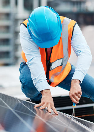 Mann in Arbeitsschutzkleidung hockt auf einem Dach und befestigt Solarpanele.