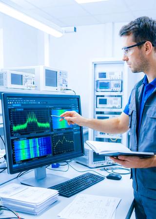 Ingenieur analysiert Funkmessdaten auf Bildschirm, daneben technische Unterlagen und Normendokumente.