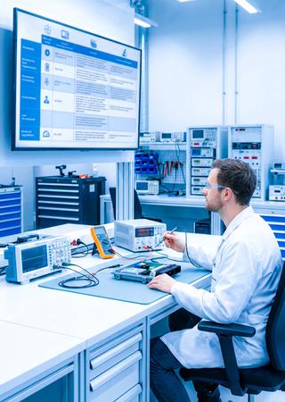  Techniker analysiert Telematik-Steuergerät am Messplatz mit Normenübersicht auf Monitor