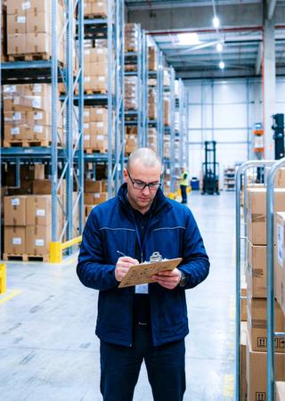 upply-Chain-/Logistik-Szene mit Containern, Lagerhalle oder Fördertechnik, dazu eine Person mit Tablet/Clipbord, die prüft oder dokumentiert 