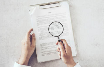 Eine Person hält ein Dokument in der einen Hand und hält mit der anderen Hand eine Lupe darüber.