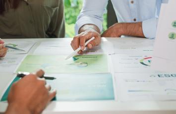 Auf einem Tisch liegen verschiedene Dokumente. Um den Tisch herum sitzen mehrere Personen. Zu sehen sind ihre Hände, die auf ein bestimmtes Blatt verweisen.
