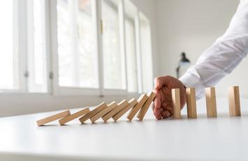 Darstellung des Dominoeffektes in Form von Bauklötzen. Etwa in der Mitte der Reihe sorgt eine Hand dafür, dass die anderen Bausteine geschützt bleiben.
