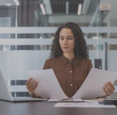 Frau sitzt am Schreibtisch und hält in den Händen zwei Dokumente, die sie betrachtet.