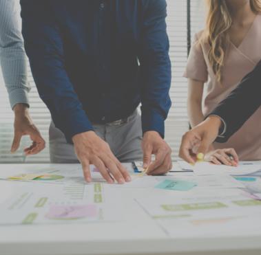 Im Vordergrund ist ein Schreibtisch mit verschiedenen Dokumenten zu sehen. Dahinter befinden sich Personen, die auf diese zeigen.