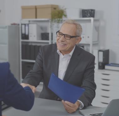 Zwei Männer sitzen sich gegenüber. Einer der beiden hält eine Dokumentenmappe in der Hand und schüttelt dem anderen die Hand.