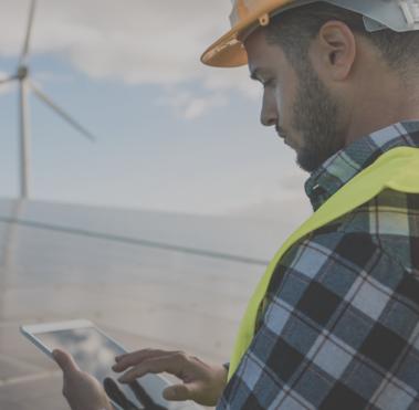 Seitenprofil eines Mannes, der Arbeitsschutzkleidung trägt und ein Tablet in der Hand hält. Im Hintergrund sind Solarpanele und Windkrafträder zu sehen.